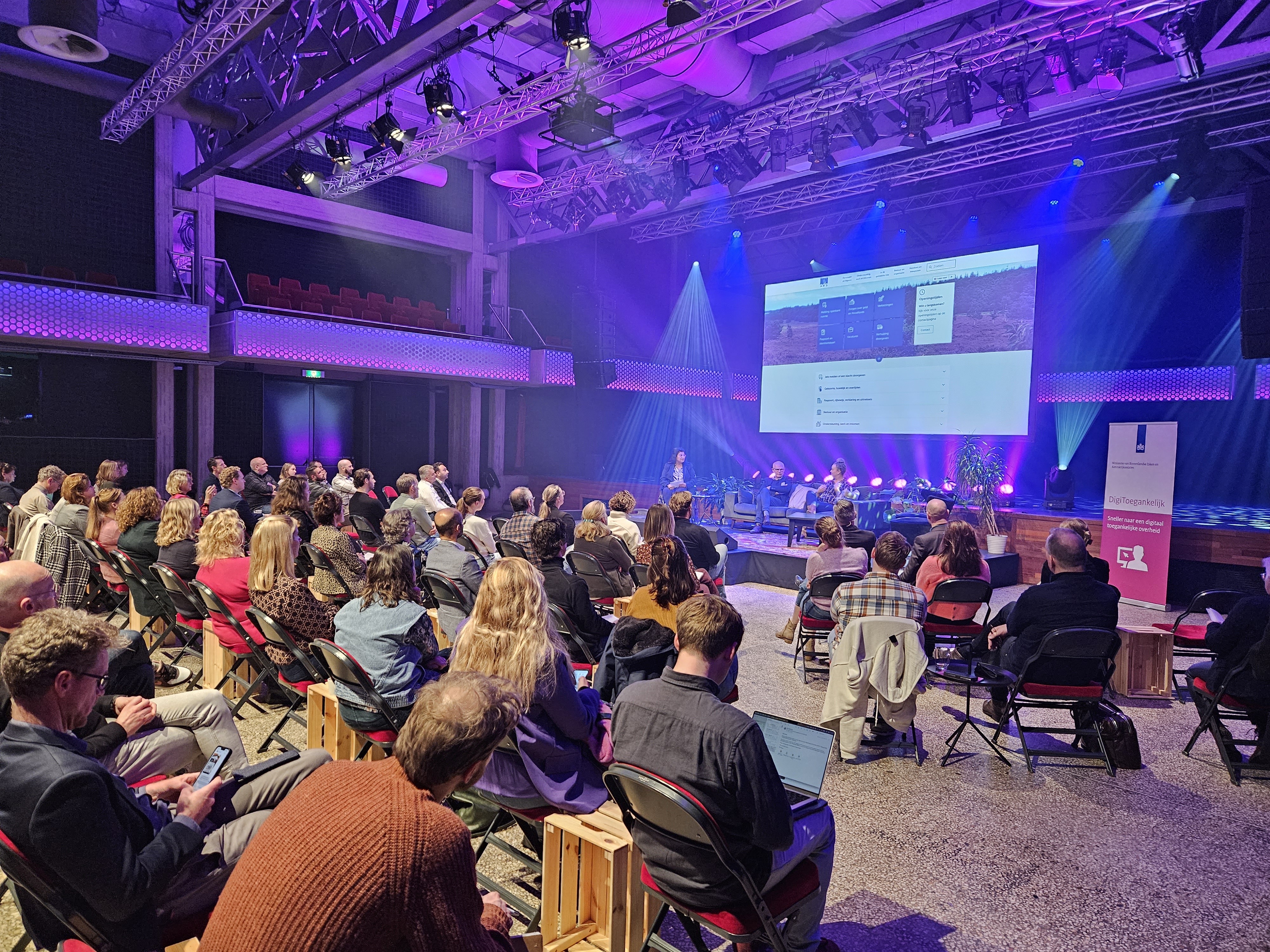 Een volle theaterzaal met op het podium presentator en sprekers, en achter het podium een groot scherm