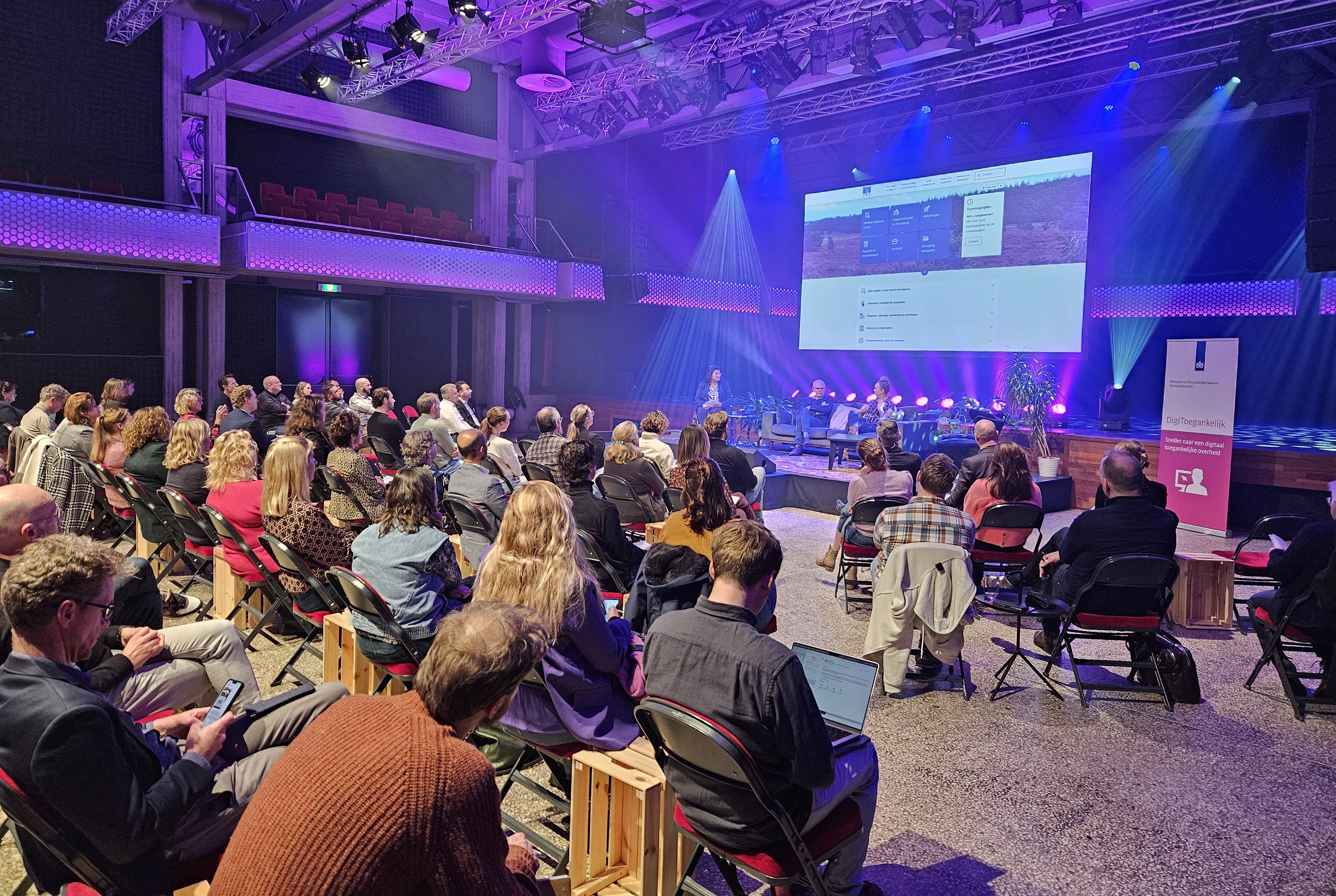 Een volle theaterzaal met op het podium presentator en sprekers, en achter het podium een groot scherm