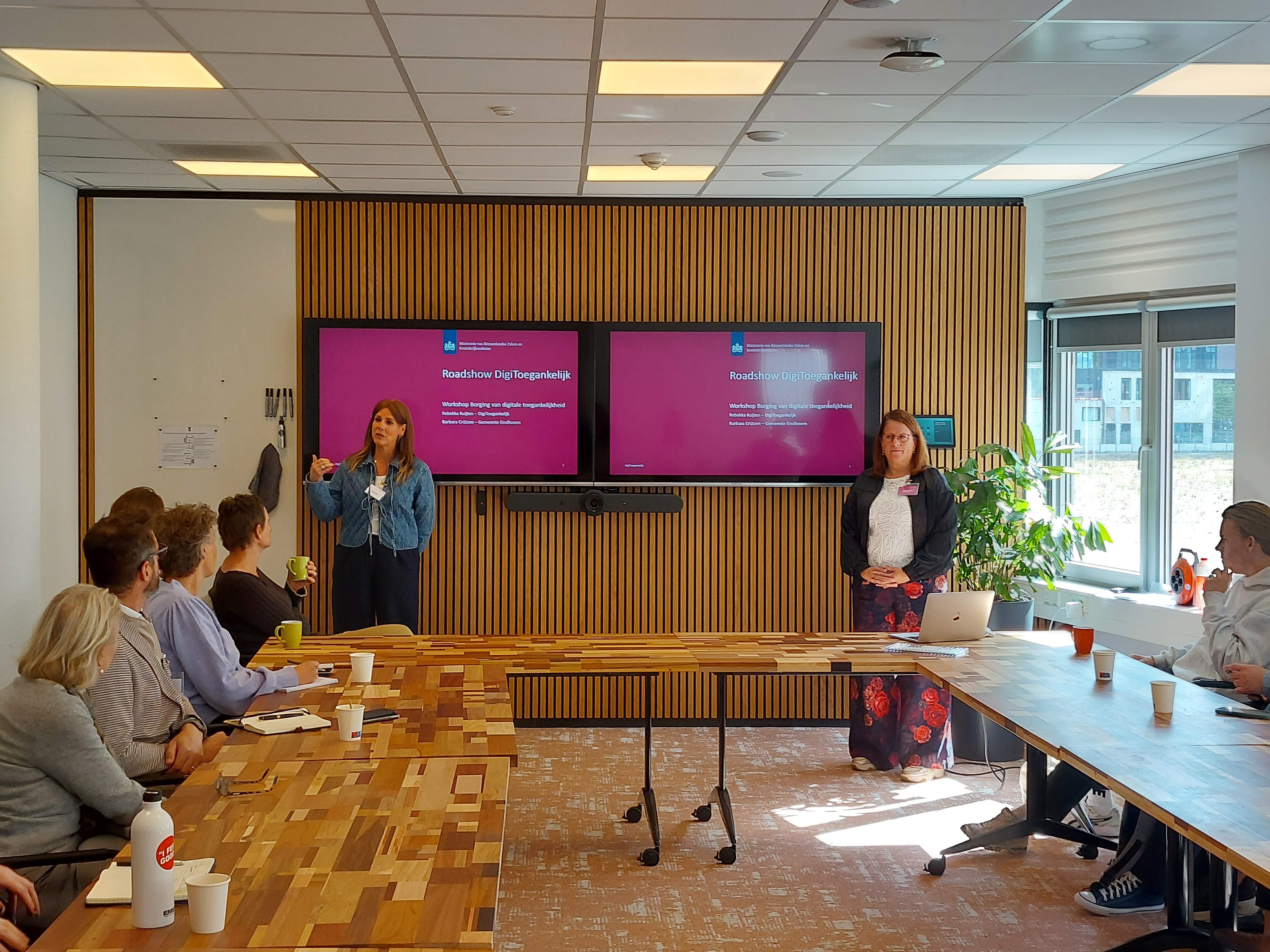 Barbara staat links voor twee presentatieschermen met rechts naast haar Rebekka. Zij presenteren voor een groep deelnemers die aan tafels in carré-vorm zitten.