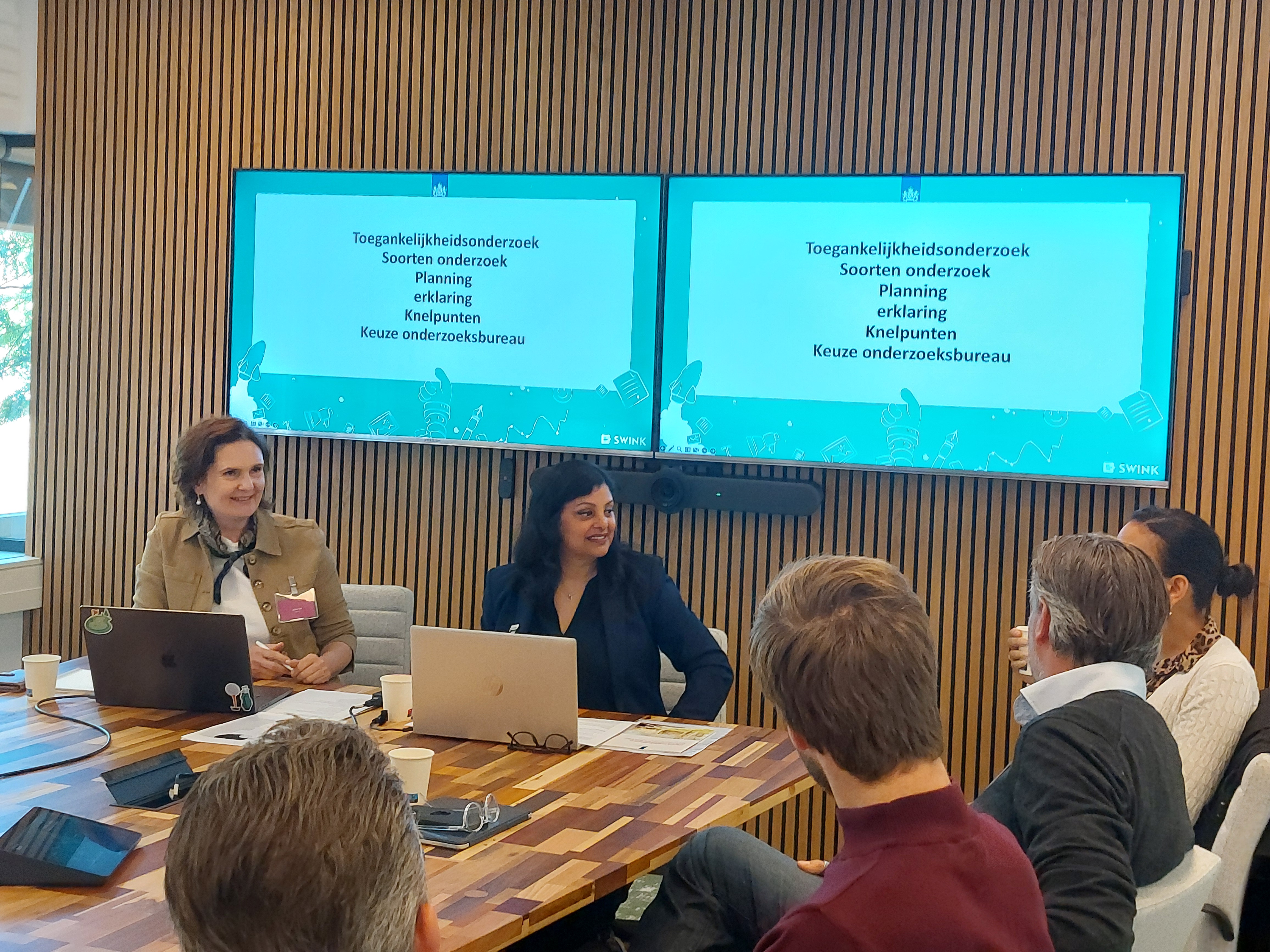Julia zit links naast Sharmila voor twee presentatieschermen aan de kop van de tafel met om hen heen de deelnemers van de workshop