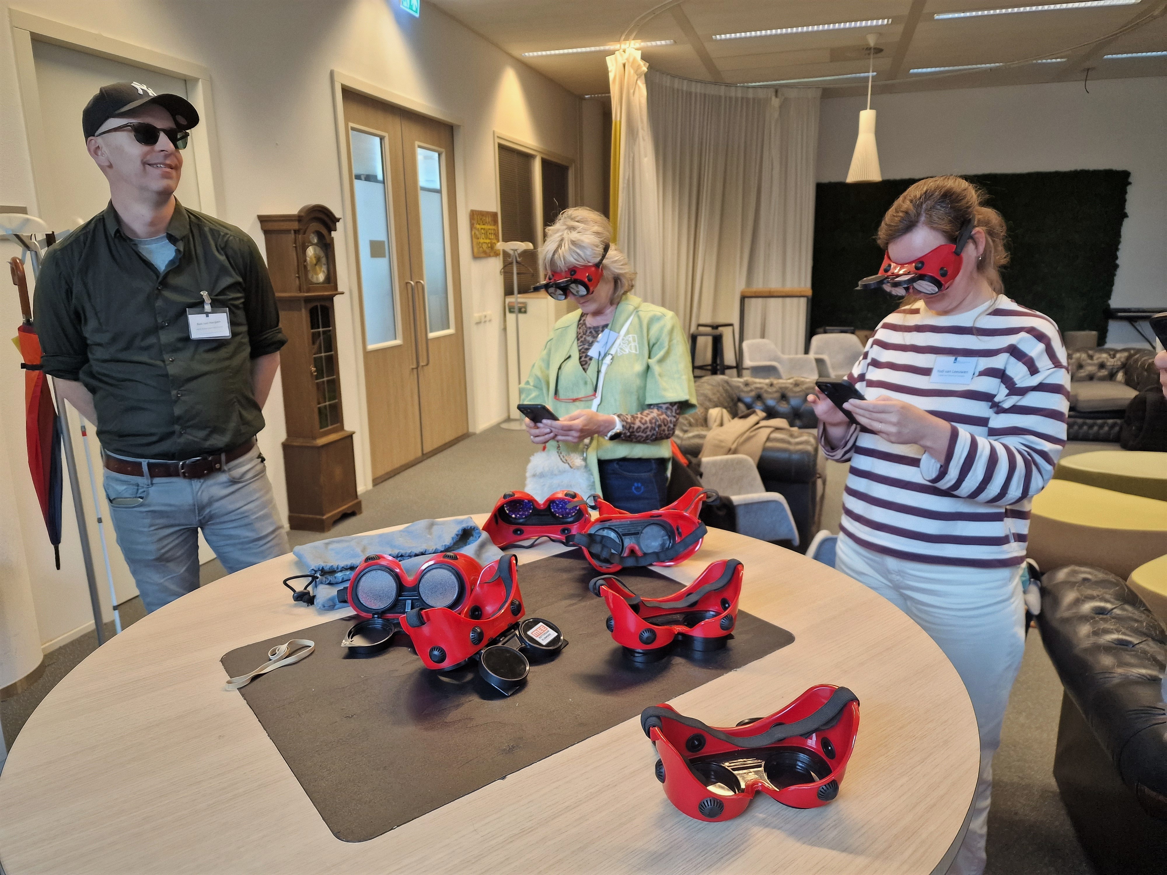 Rondom een tafel met simulatiebrillen voor visuele beperkingen staan drie personen. Twee van hen dragen de bril en kijken op hun telefoon.