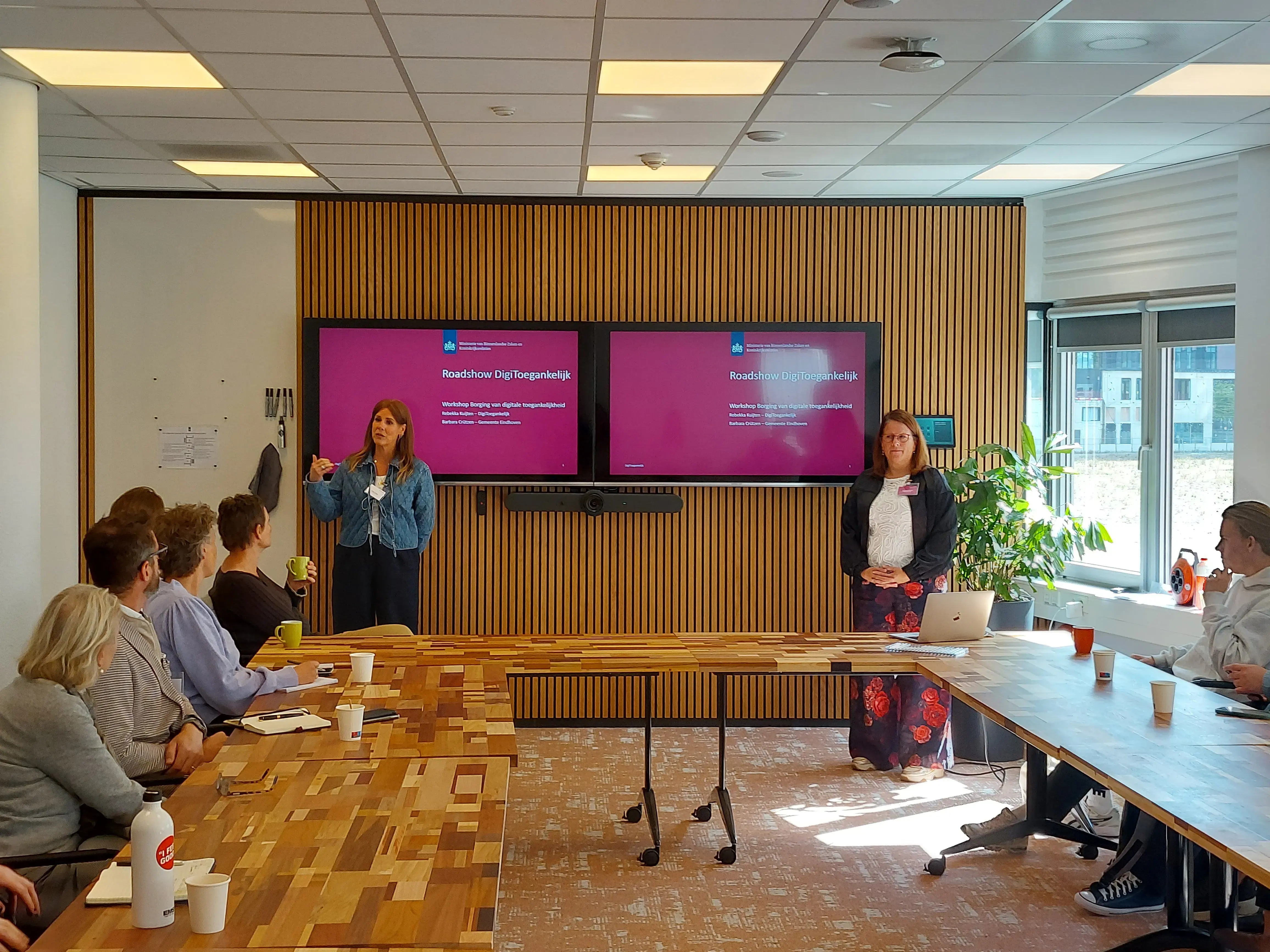 Barbara staat links voor twee presentatieschermen met rechts naast haar Rebekka. Zij presenteren voor een groep deelnemers die aan tafels in carré-vorm zitten.