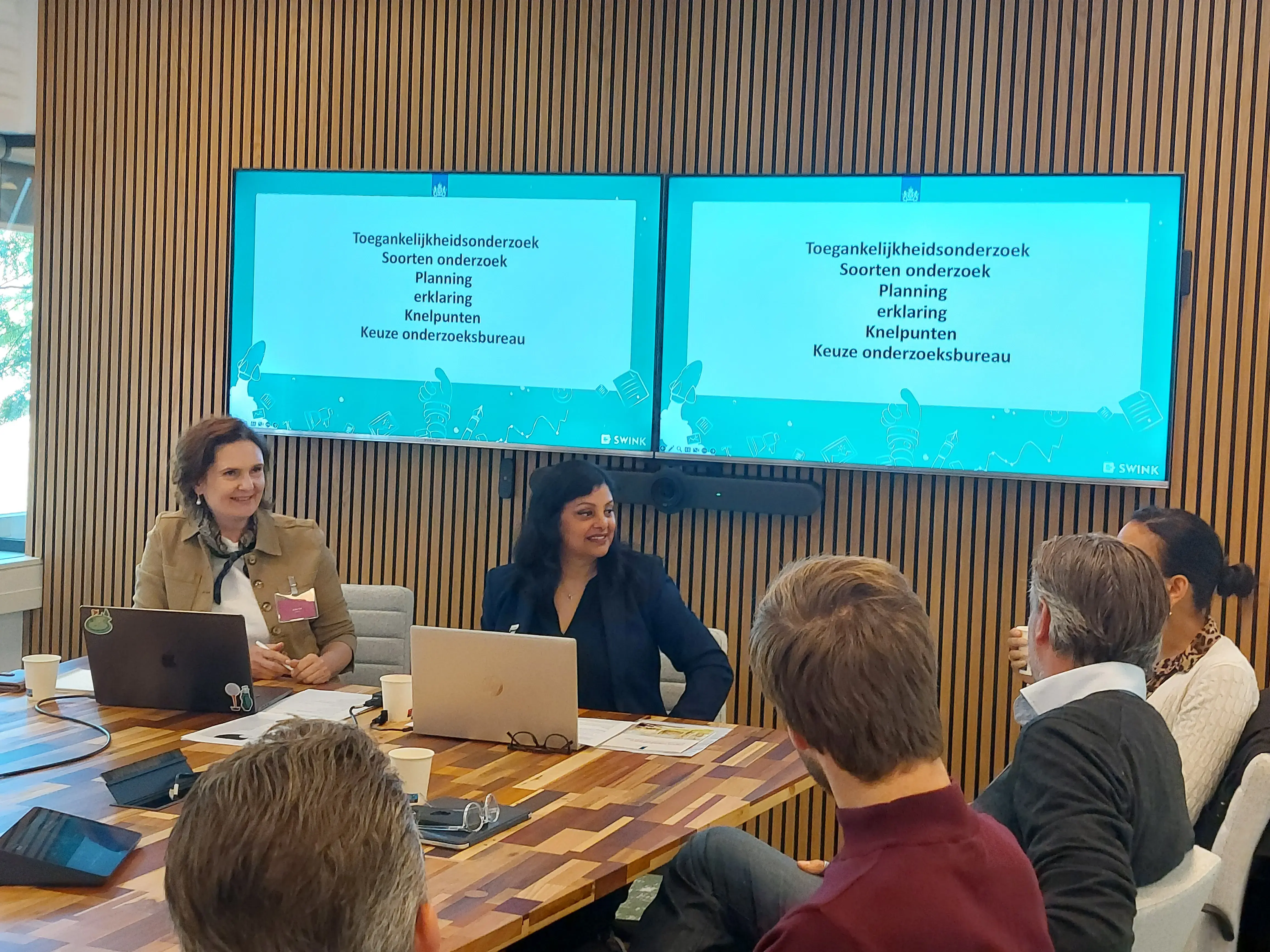 Julia  zit links naast Sharmila voor twee presentatieschermen aan de kop van de tafel met om hen heen de deelnemers van de workshop